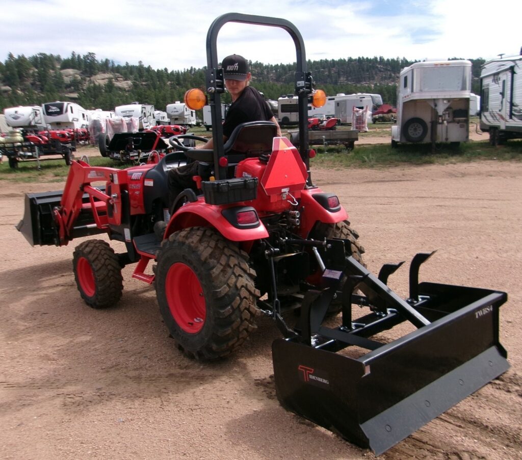 Man sitting on 24HP KIOTI Compact Tractor and Rear Box Blade in dirt parking lot